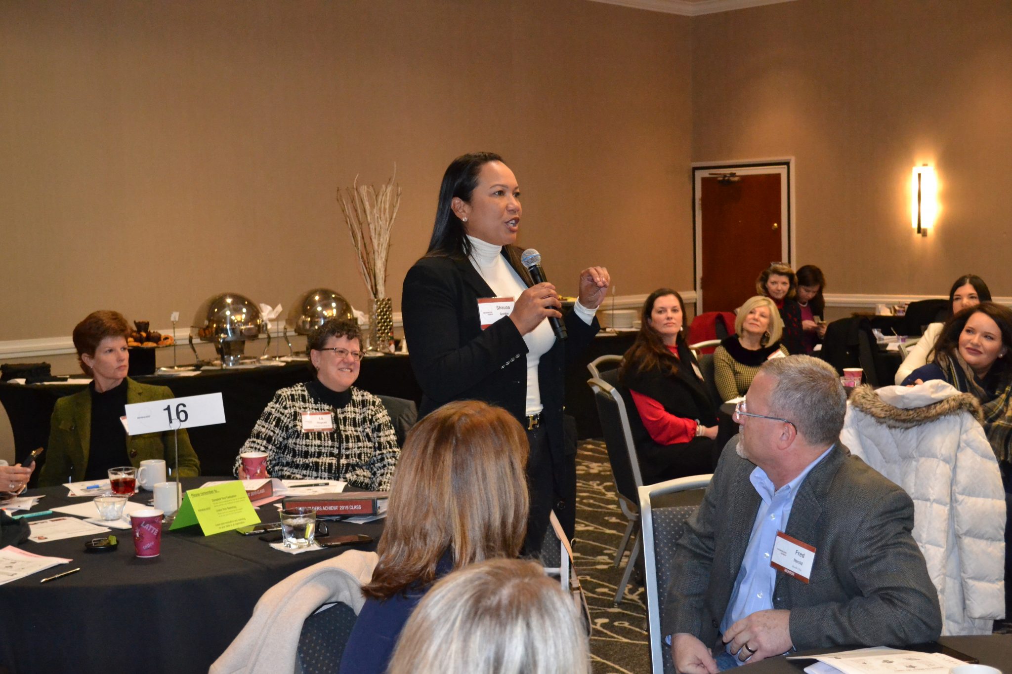 A leader speaking in a Pathbuilders session — seen first through a narrow frame on the speaker, then widened to reveal the room of people listening.